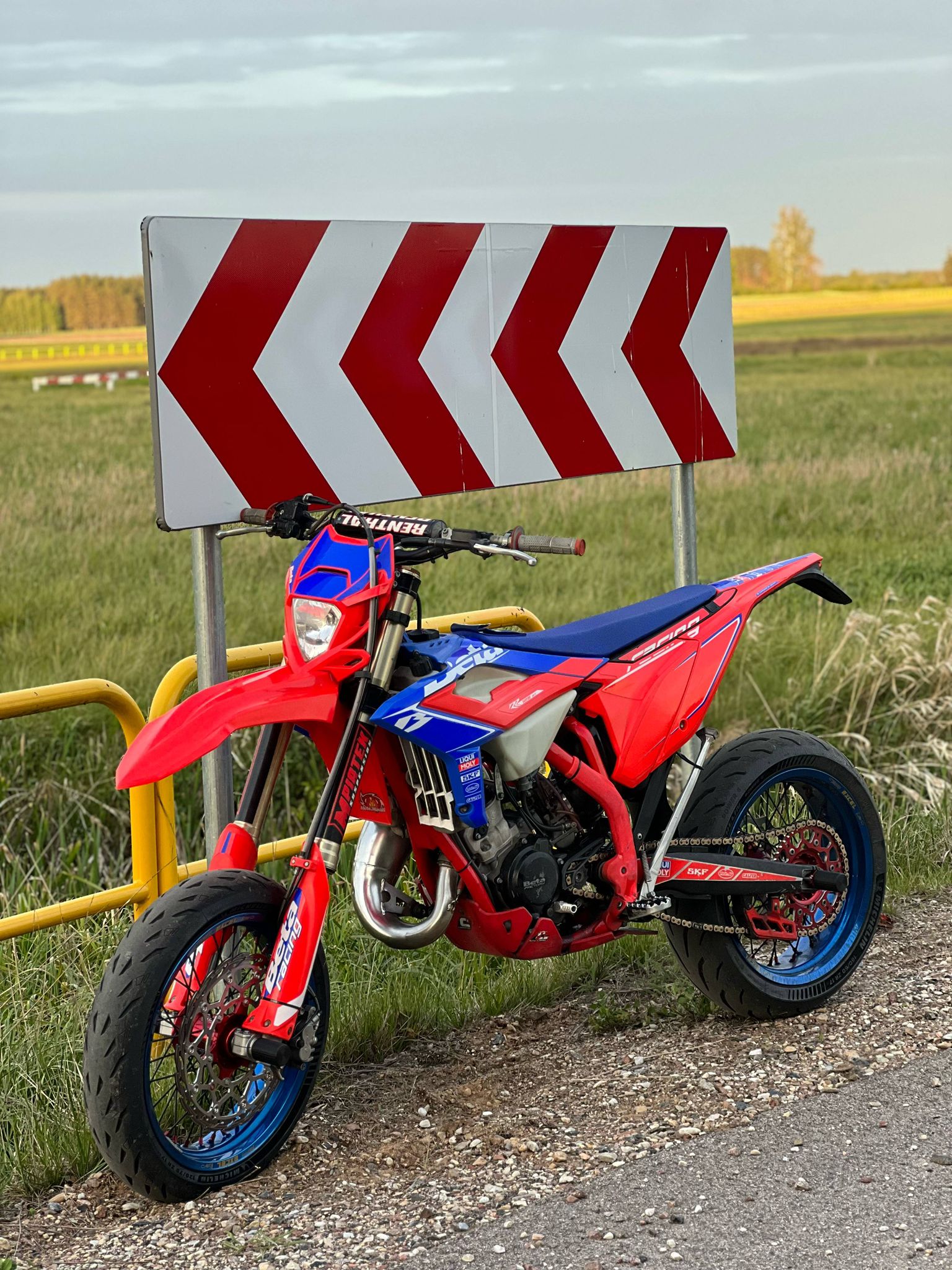 Red and blue dirt bike parked on a road next to a warning sign with a yellow field in the background.