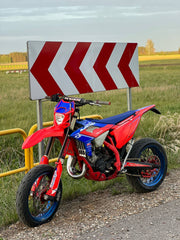 Red and blue dirt bike parked on a road next to a warning sign with a yellow field in the background.